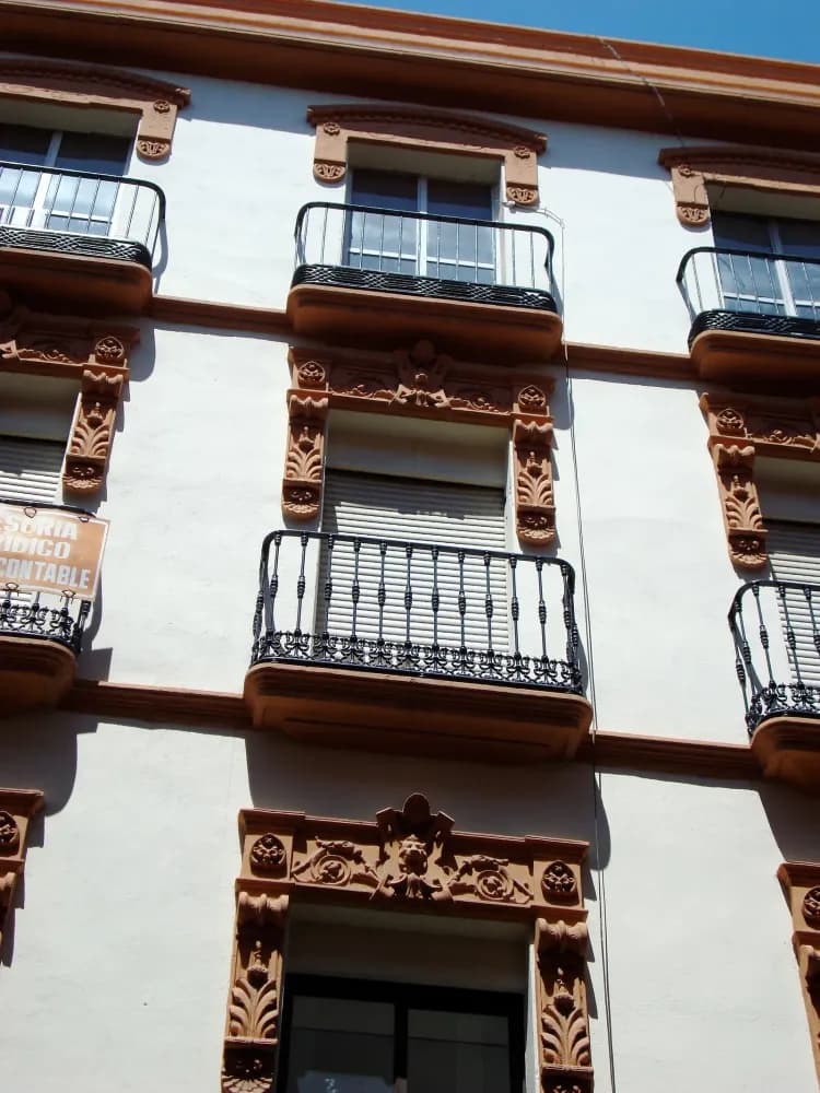 Facade of a white Spanish apartment building with ornate terracotta trim, wrought-iron balconies, and a partial street sign.